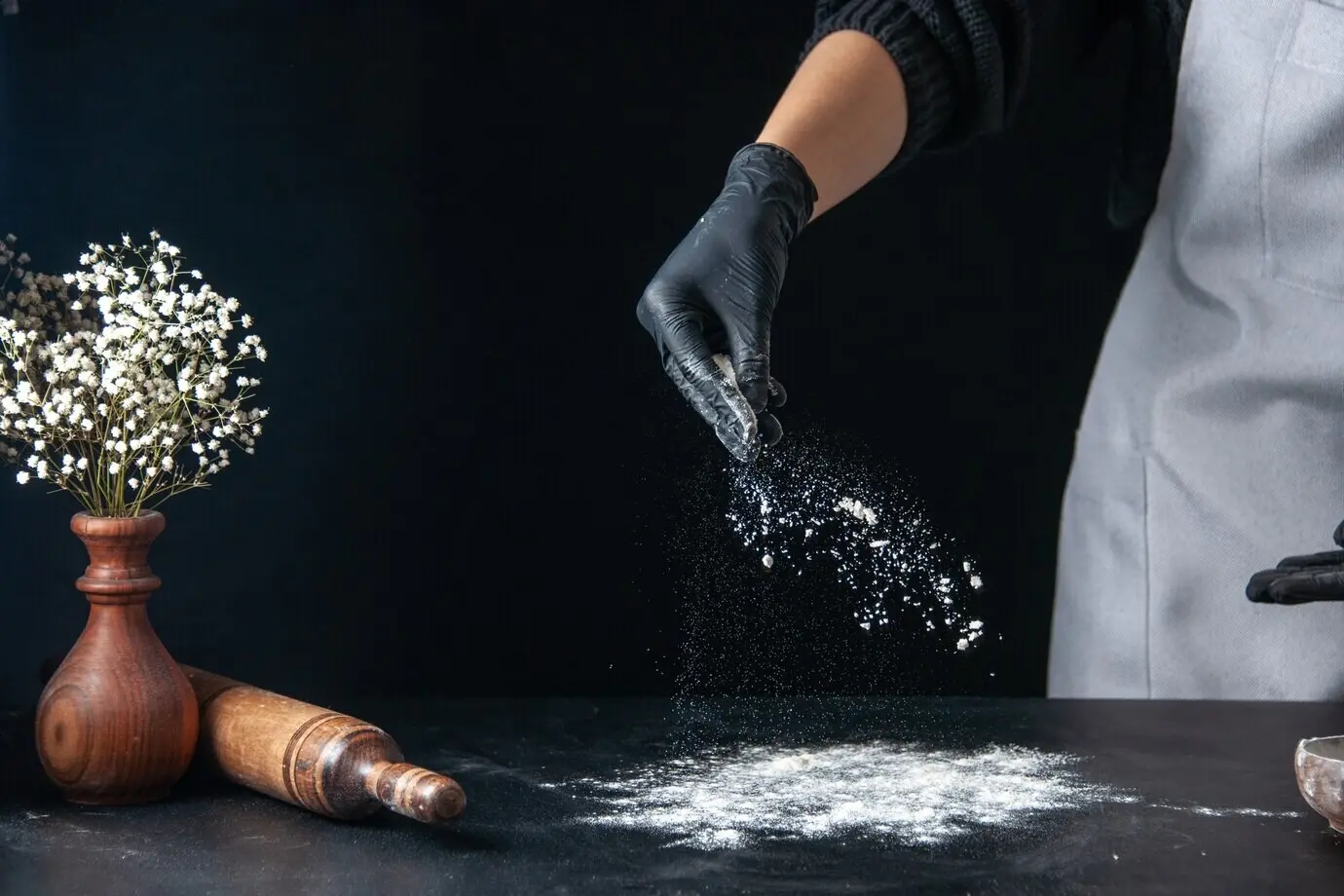 Vorderansicht einer Köchin, die in einer dunklen Konditorei-/Bäckereiküche Mehl für den Teig auf den Tisch schüttet; Ei, Küchenarbeit, Pfannkuchen.